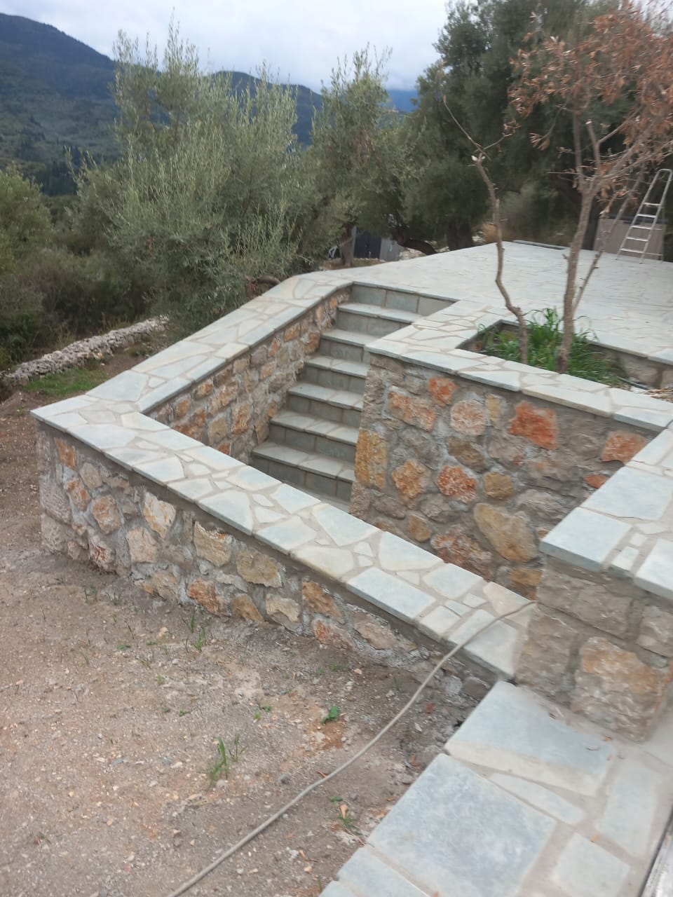 Stone and concrete staircase with surrounding stone walls, outdoors with trees and mountains in background.