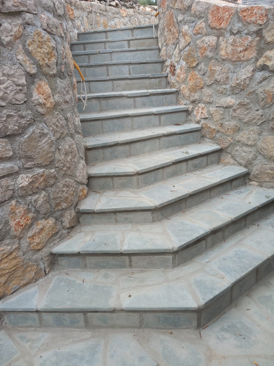 Stone stairs with metal handrail between stone walls, ascending upward.