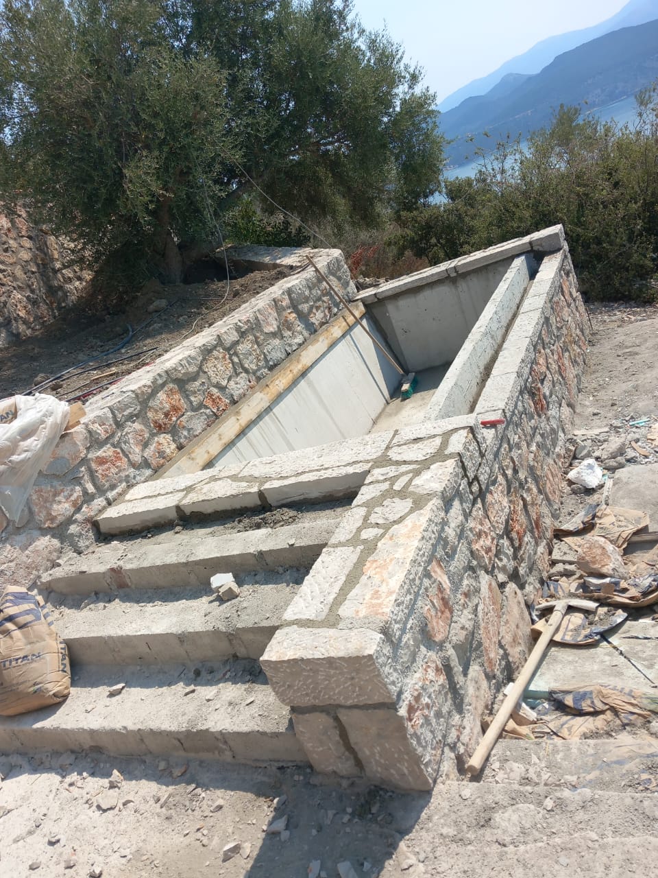 Stone and concrete structure under construction with stairs and a sloped wall, outdoors with trees and mountains in background.