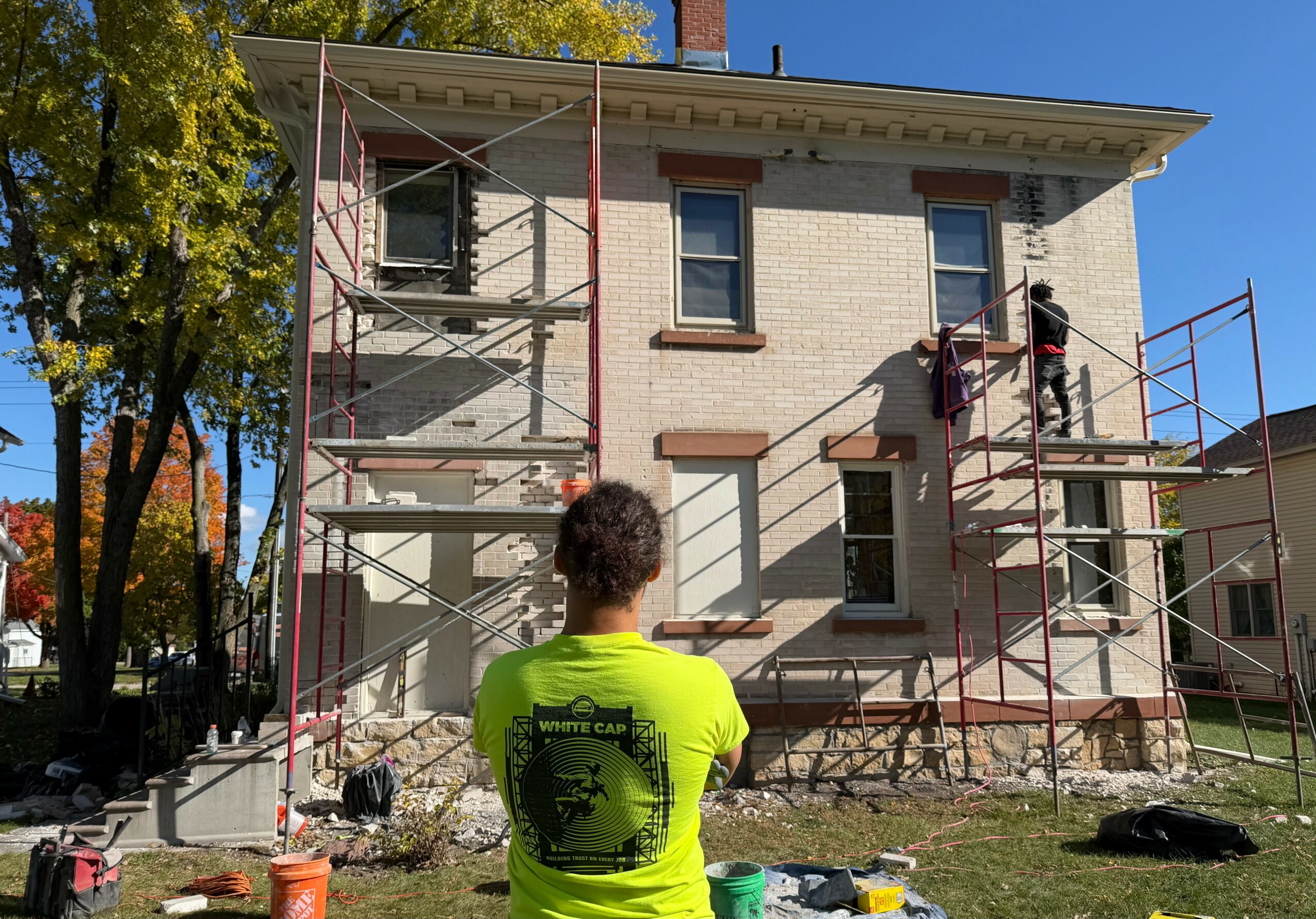 Person in yellow shirt observing a house renovation with scaffolding and two workers on the building's exterior.