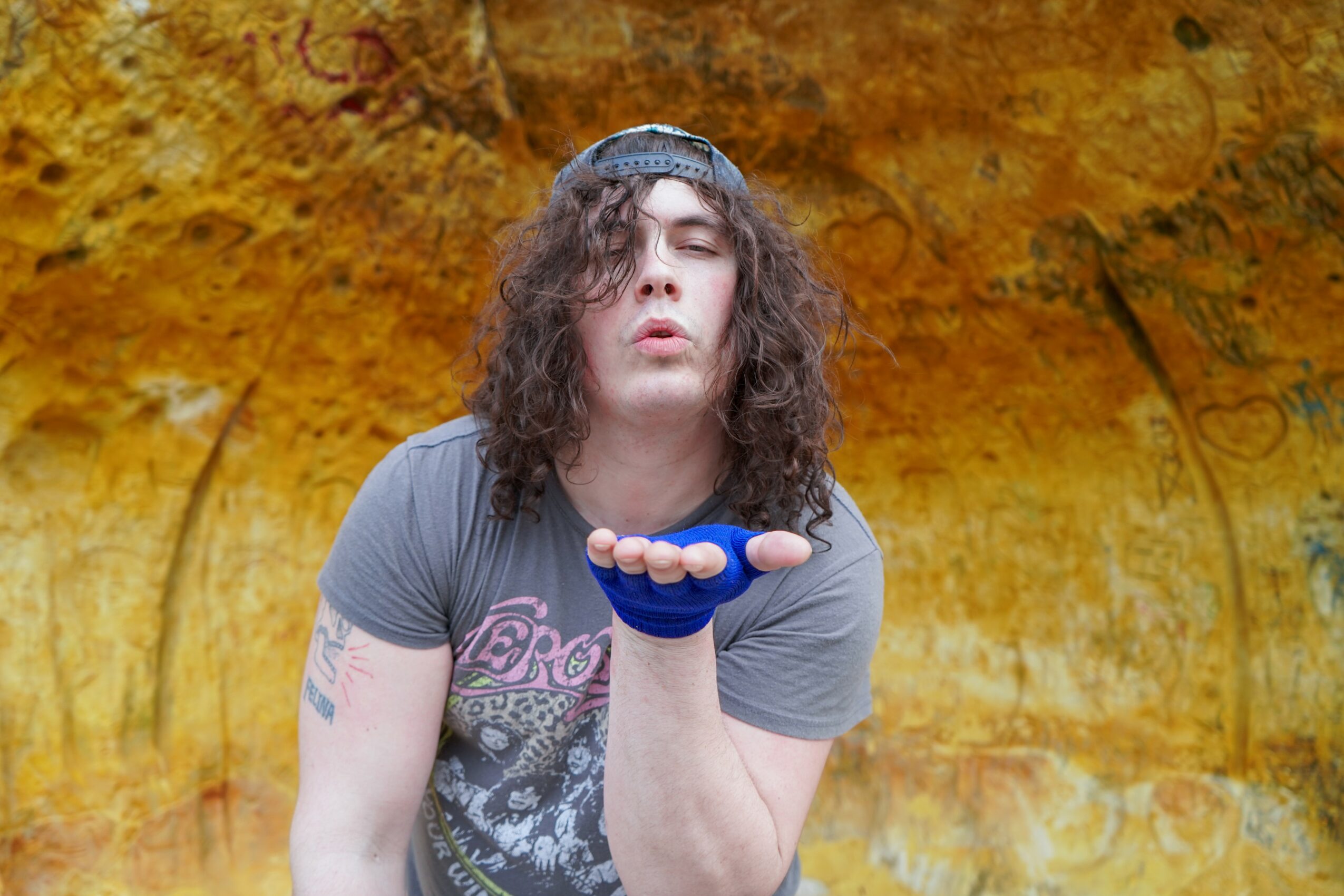Person with long curly hair wearing a gray T-shirt and a headband, holding hand near face, standing outdoors.