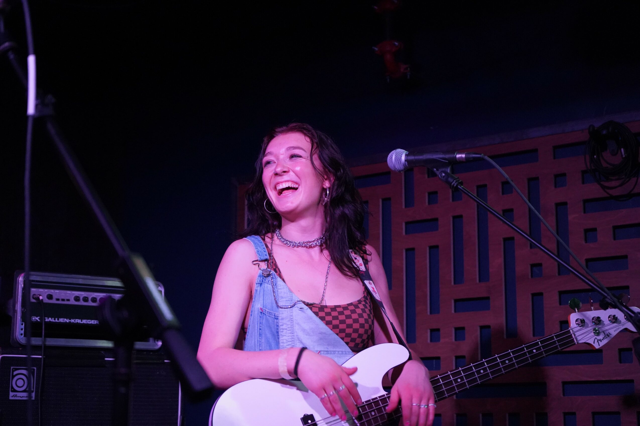 Young woman with long dark hair smiling, holding a guitar, on stage with microphone and sound equipment.