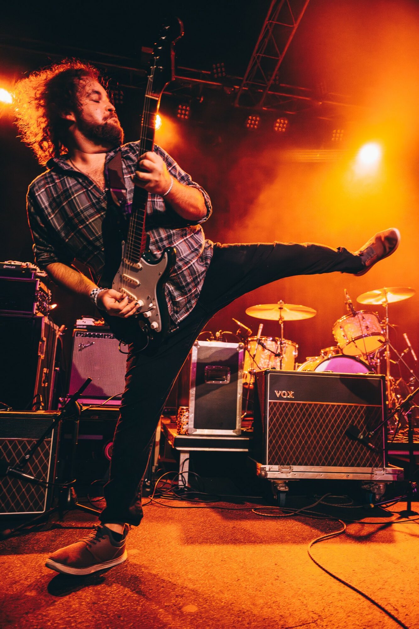 Musician with curly hair playing electric guitar on stage with drums and amplifiers, colorful stage lighting.