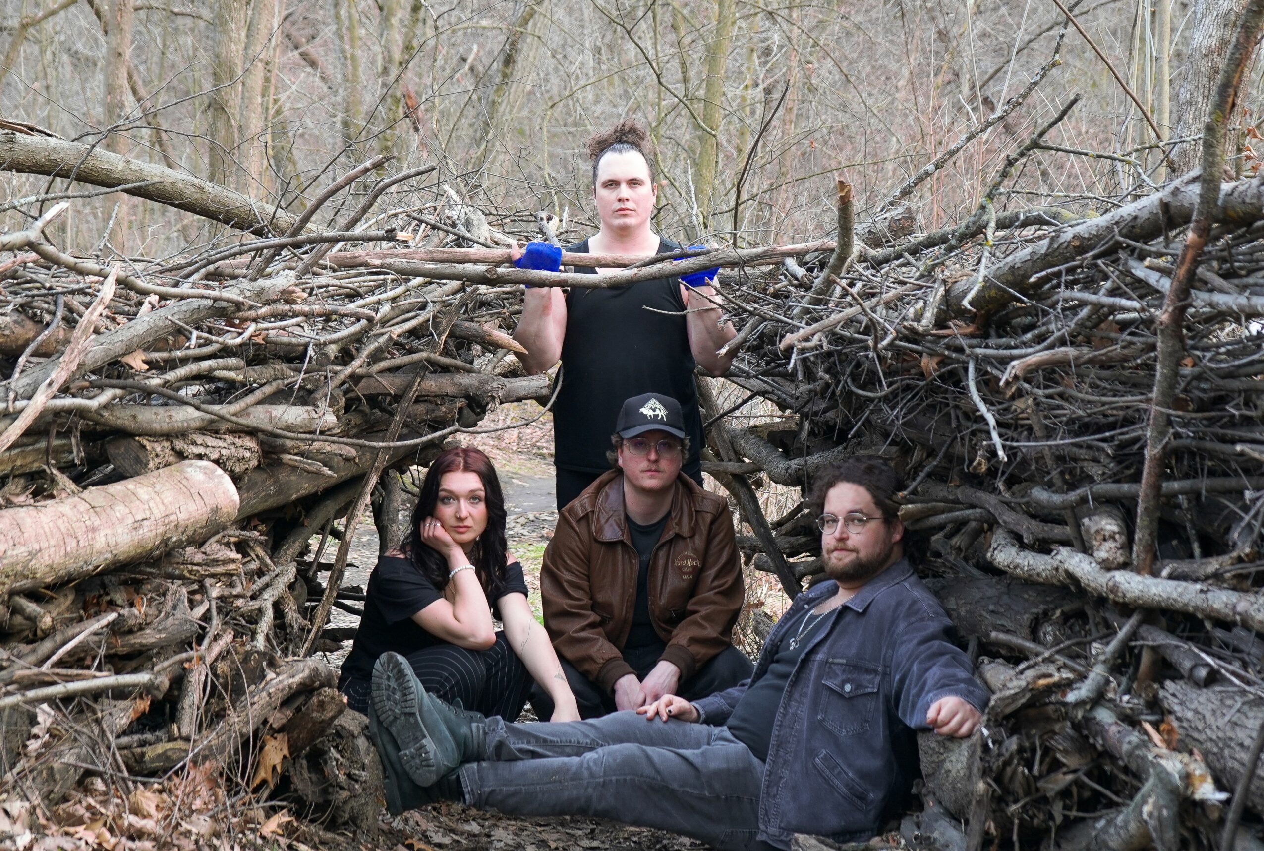 Four people sit among fallen branches in a wooded area, with one person standing behind holding a large branch structure.