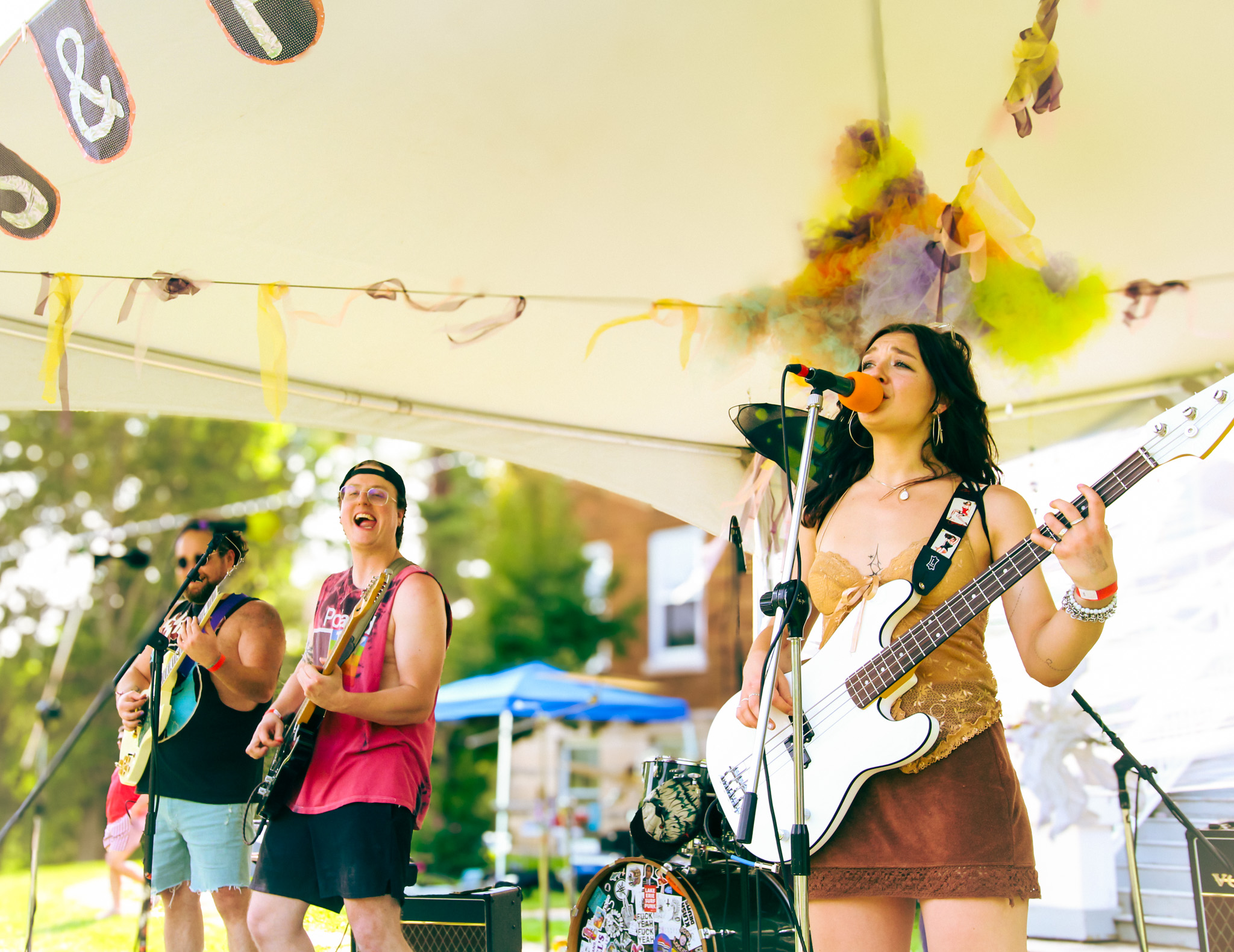 Three musicians perform outdoors under a decorated canopy, with trees and buildings in the background.