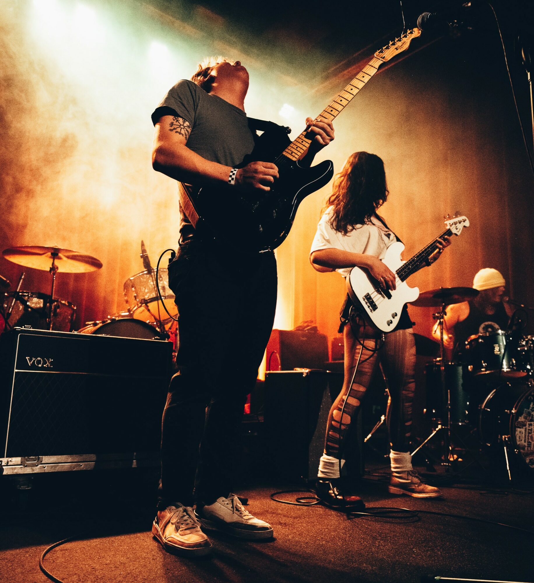 Musicians perform on stage with guitars, surrounded by equipment and warm lighting.