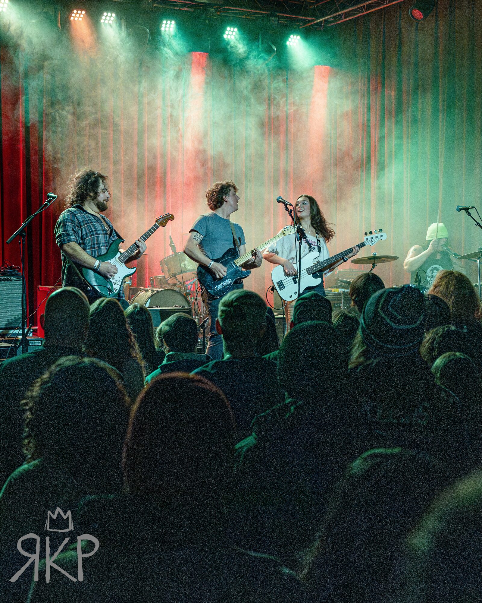 Band performing on stage with three guitarists and a drummer, audience watching in a dimly lit venue with colorful lights.