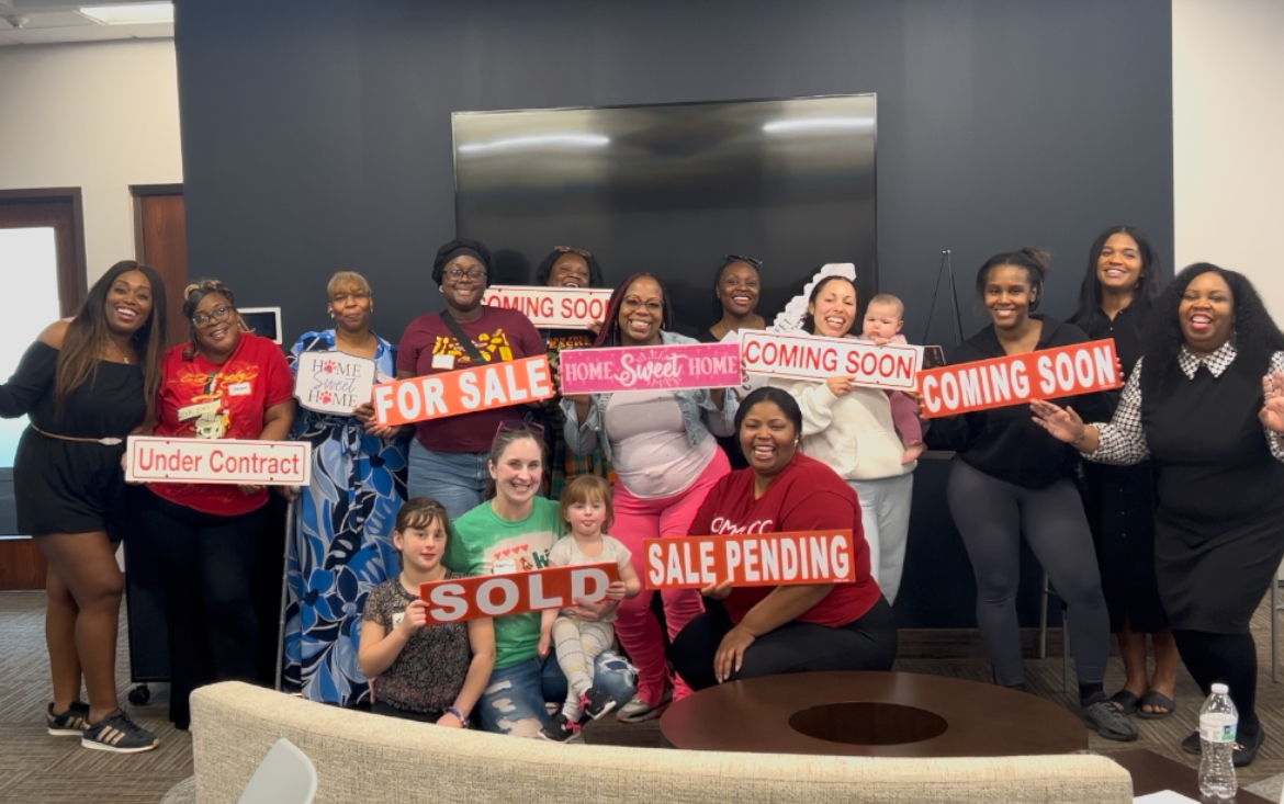 Group of people holding signs, some with 'Sold', 'For Sale', and 'Coming Soon' messages, in a room with a large screen.