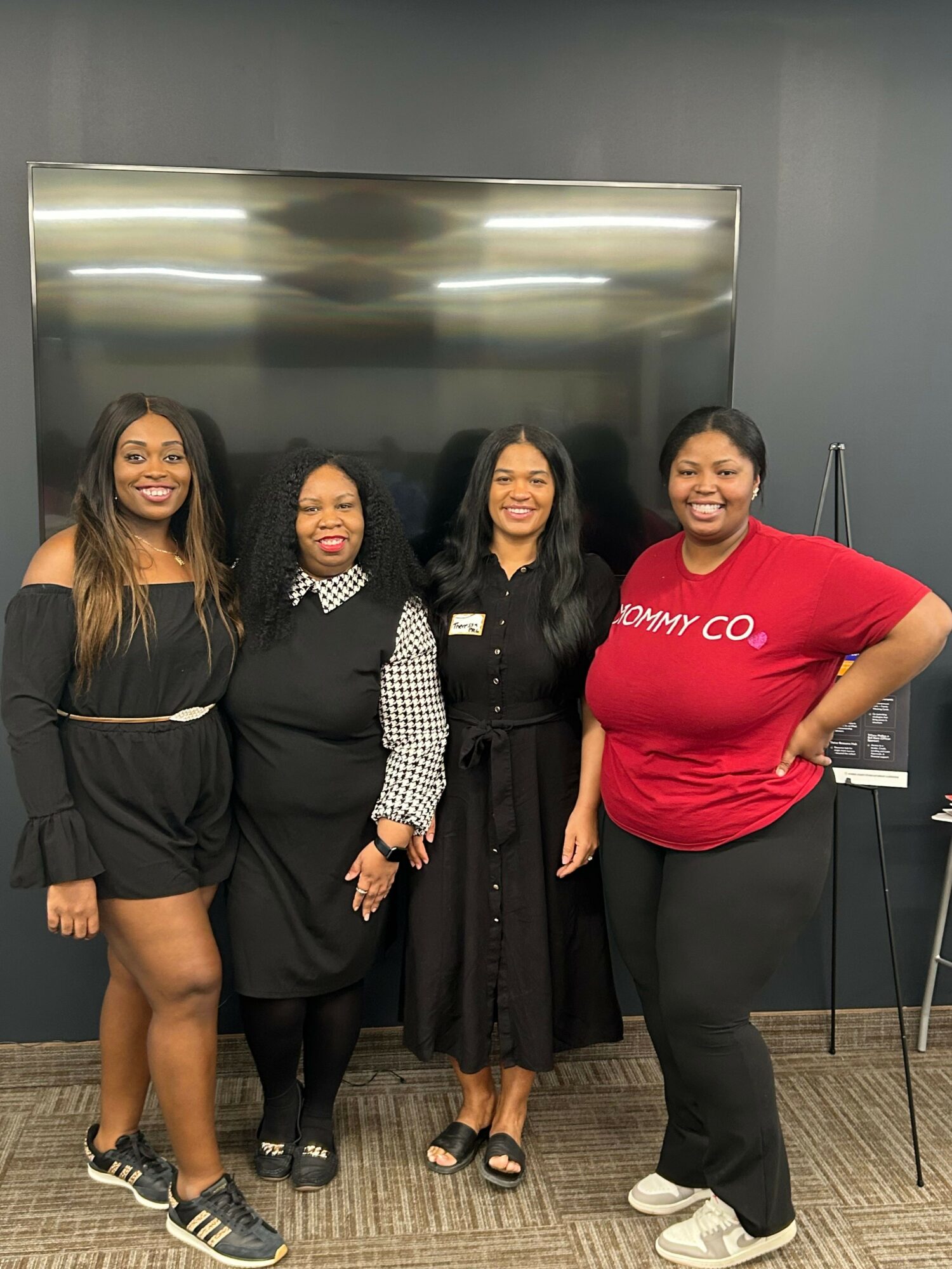 Four women standing together indoors, smiling, in front of a black wall and metallic panel.