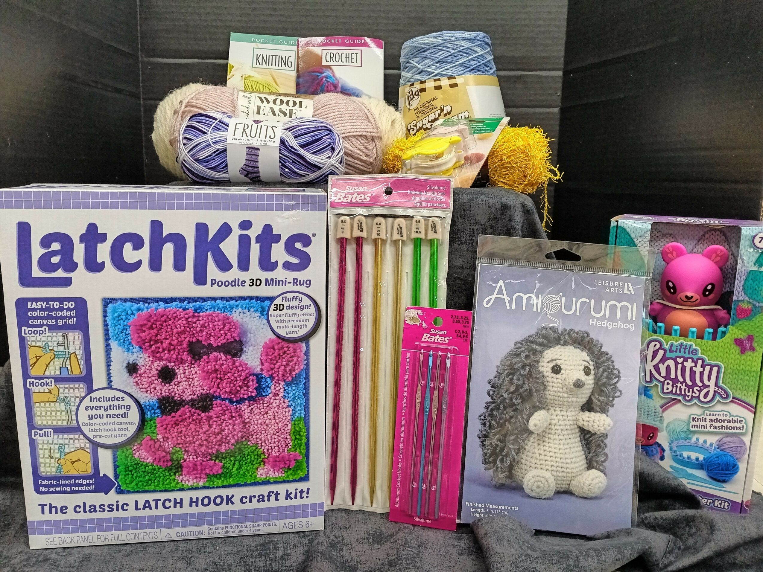 Various knitting and crochet supplies, including yarn, latch hook kit, knitting needles, and a plush doll, arranged on a surface.