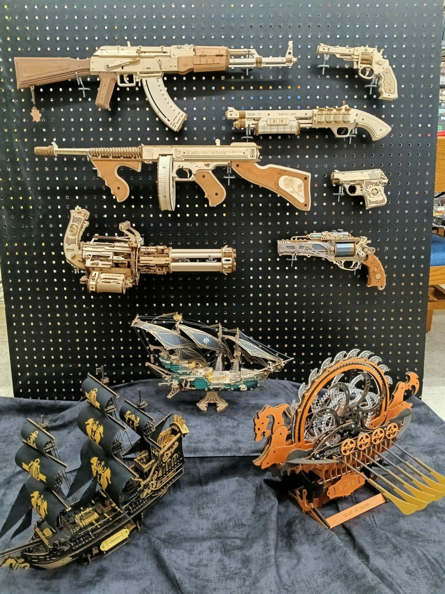 Collection of gun models and a mechanical device on a black pegboard, with a blue fabric background.