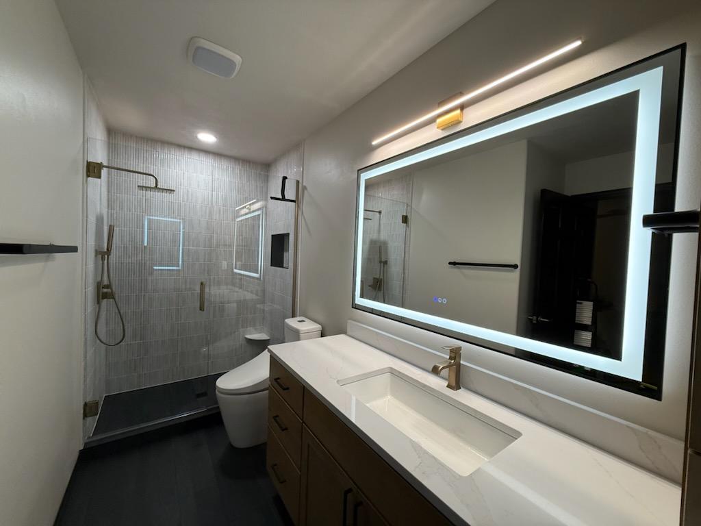 Bathroom with shower area, toilet, large mirror with light, and vanity with sink and faucet.