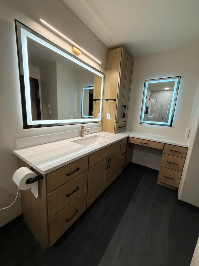 Bathroom vanity with large illuminated mirror, sink, wooden cabinets, and a window with backlit frame.