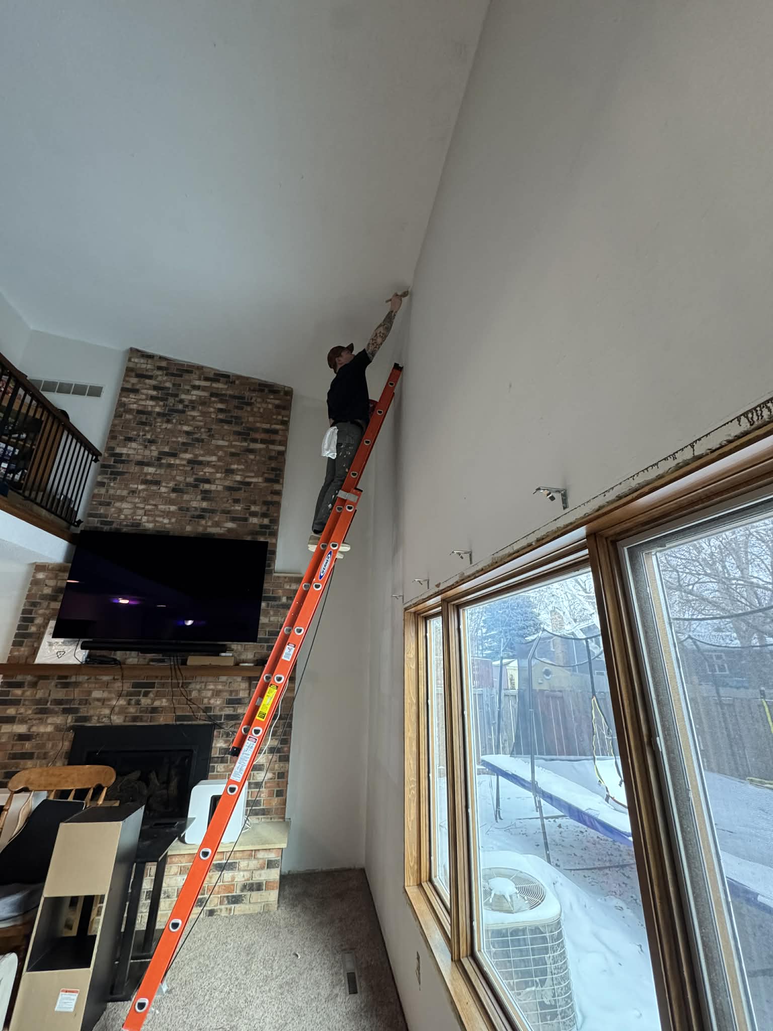 Person on an orange ladder cleaning or fixing the ceiling in a living room with large windows and a brick fireplace.