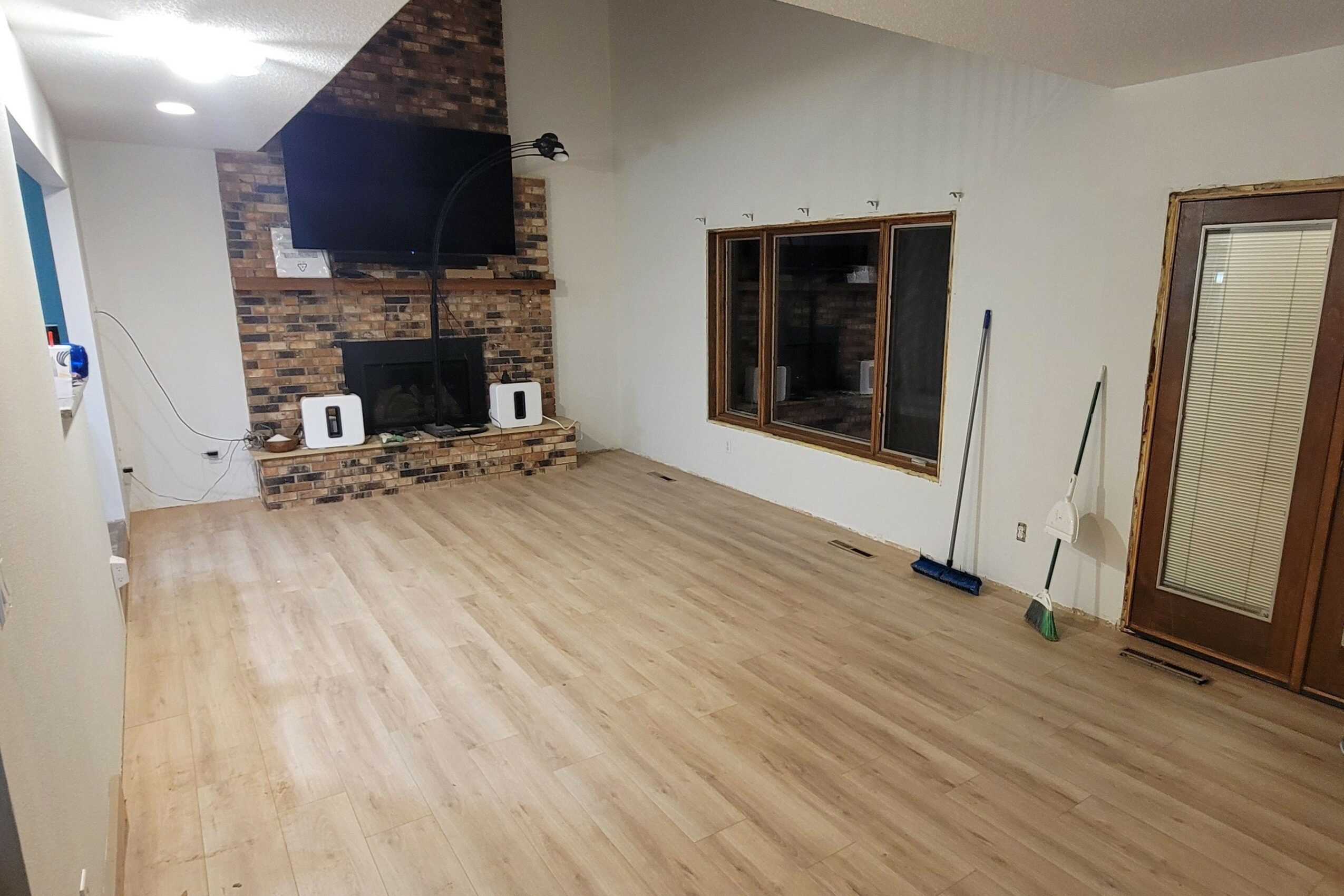 Empty living room with wooden floor, brick fireplace, wall-mounted TV, and window with a broom and cleaning tools nearby.