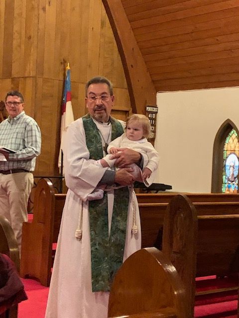 Man in religious robe holding a child inside a church with wooden walls and stained glass windows.