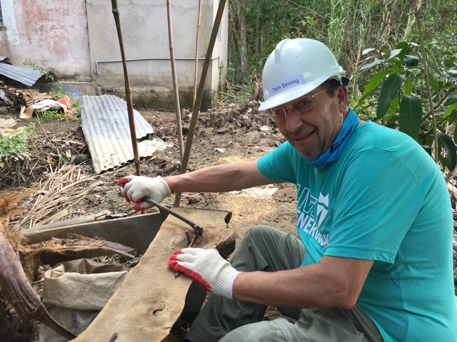 Man wearing a white helmet and blue shirt, smiling while working outdoors with tools and construction materials.