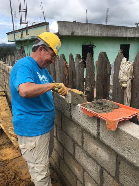 Man wearing yellow helmet and blue shirt building a brick wall outdoors, with a wooden fence and houses in background.