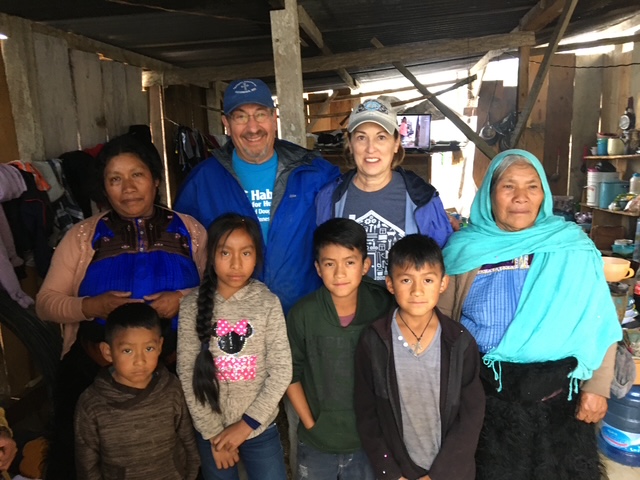 Group of seven people, three adults and four children, standing inside a rustic shelter with wooden walls and supplies.