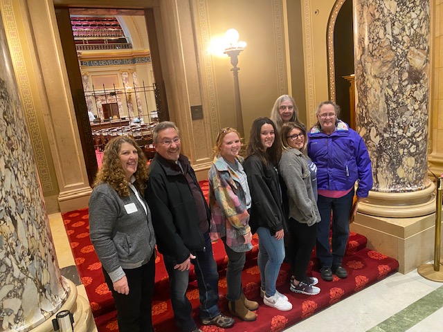 Six people standing on a red carpeted staircase in an ornate interior with marble columns and warm lighting.