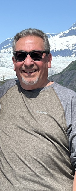 Man wearing sunglasses smiling outdoors with snow-covered mountains in background.