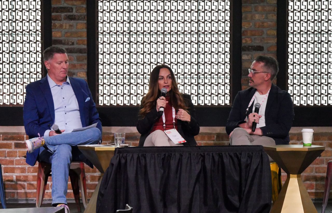 Three people seated at a panel discussion with a brick wall and patterned window behind them.