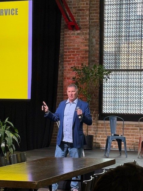 Man in blazer and jeans speaking on stage with a large screen and brick wall background.