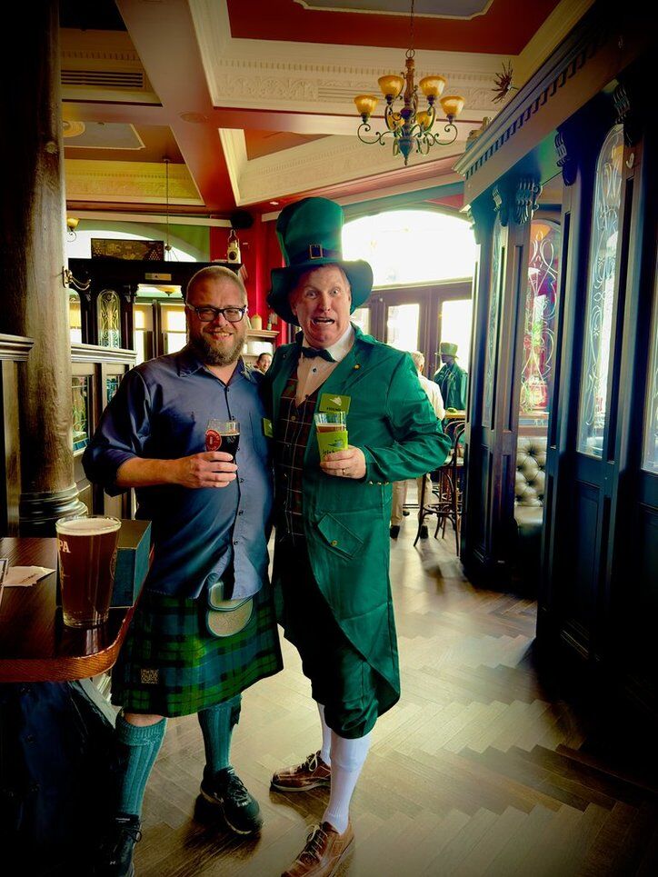 Two men in traditional attire inside a decorated pub, holding drinks and smiling.
