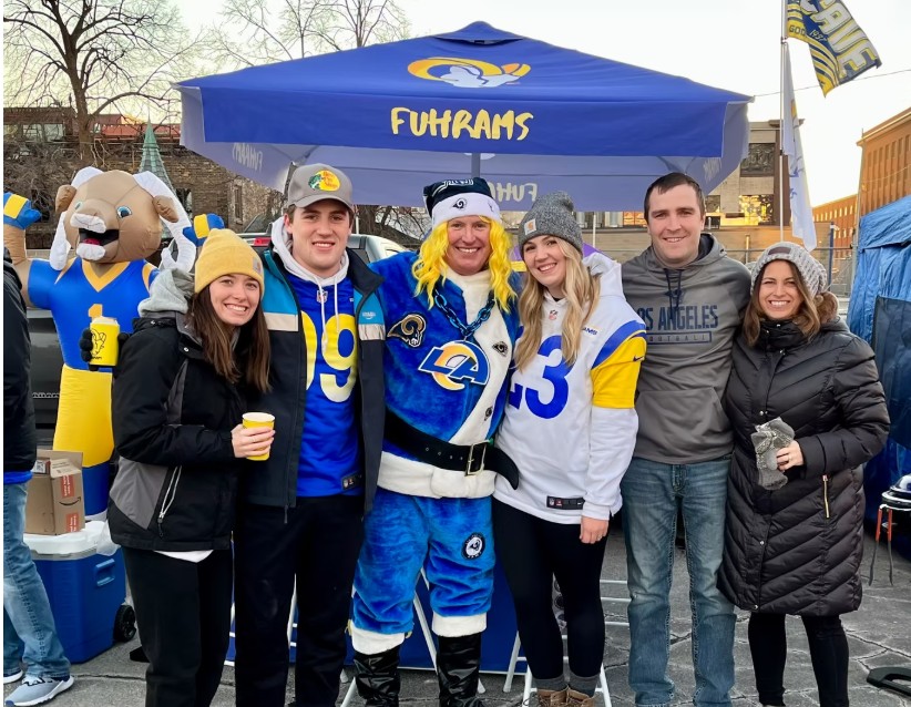 Group of five people and a mascot standing outdoors under a blue canopy, smiling, holding drinks, wearing sports jerseys and jackets.