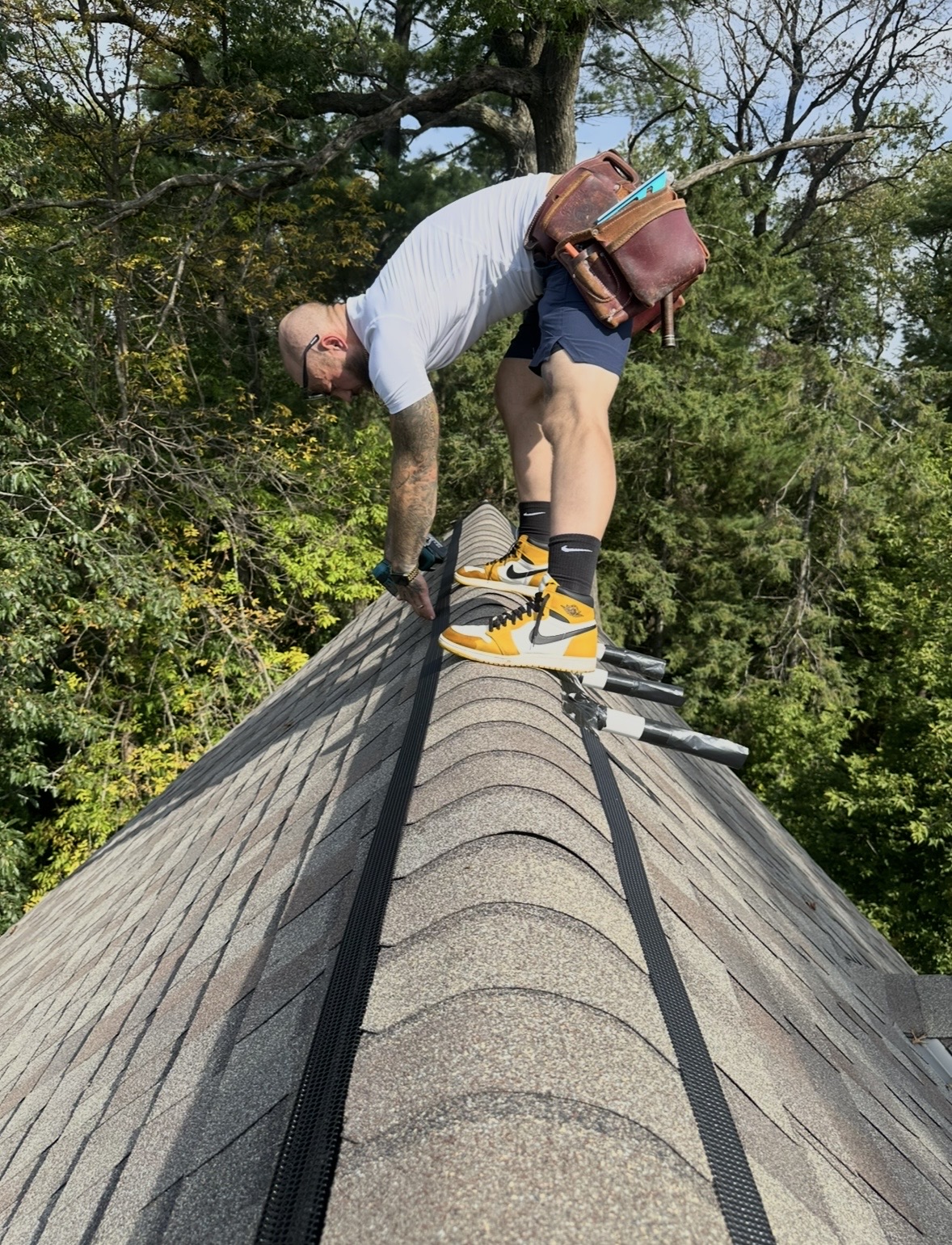 Person crouching on roof with tools, wearing shorts, a gray shirt, and yellow shoes, surrounded by trees.