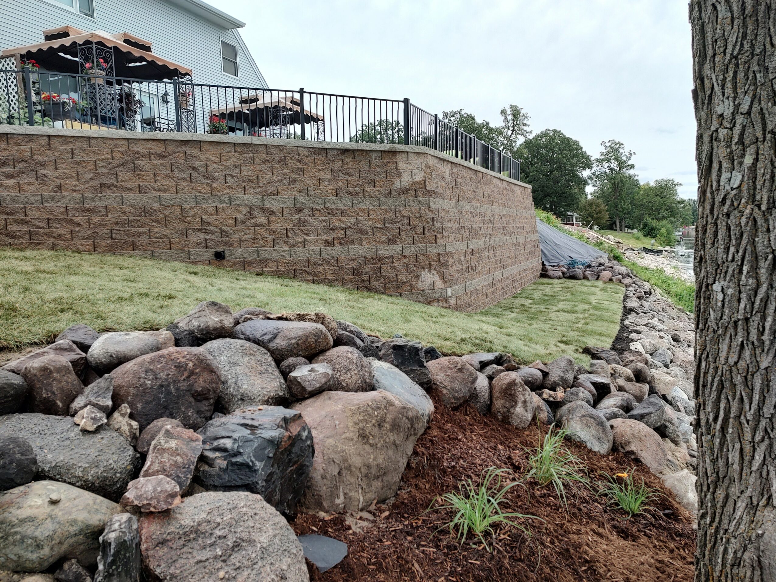 Yard with rocks, grass, and a brick retaining wall, house in background, tree trunk on right side.