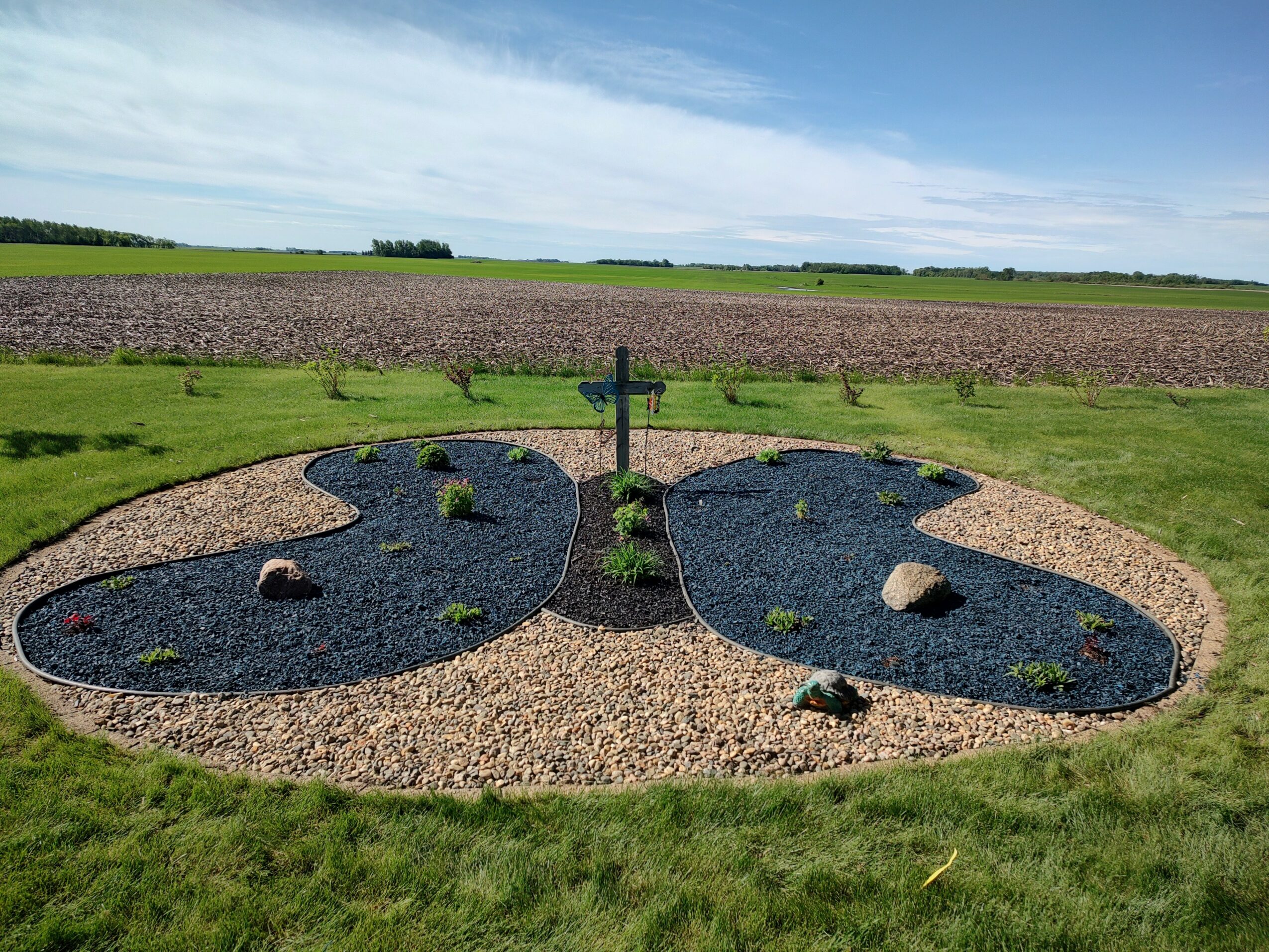 Symmetrical garden with two oval-shaped sections filled with dark gravel, surrounded by grass and a central cross-shaped plant, in a rural landscape.