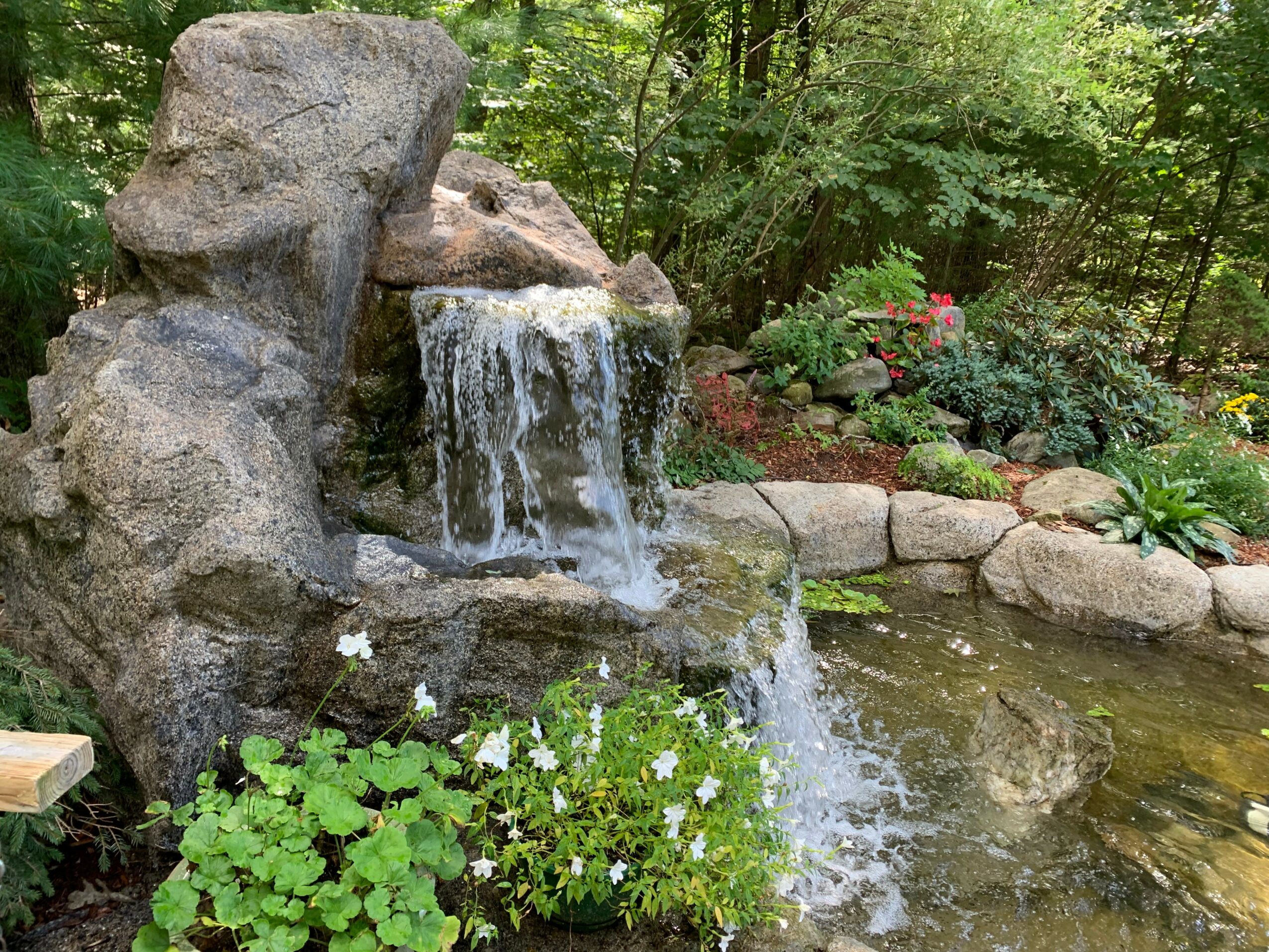 Small waterfall flowing over rocks into a pond surrounded by plants and flowers in a garden.