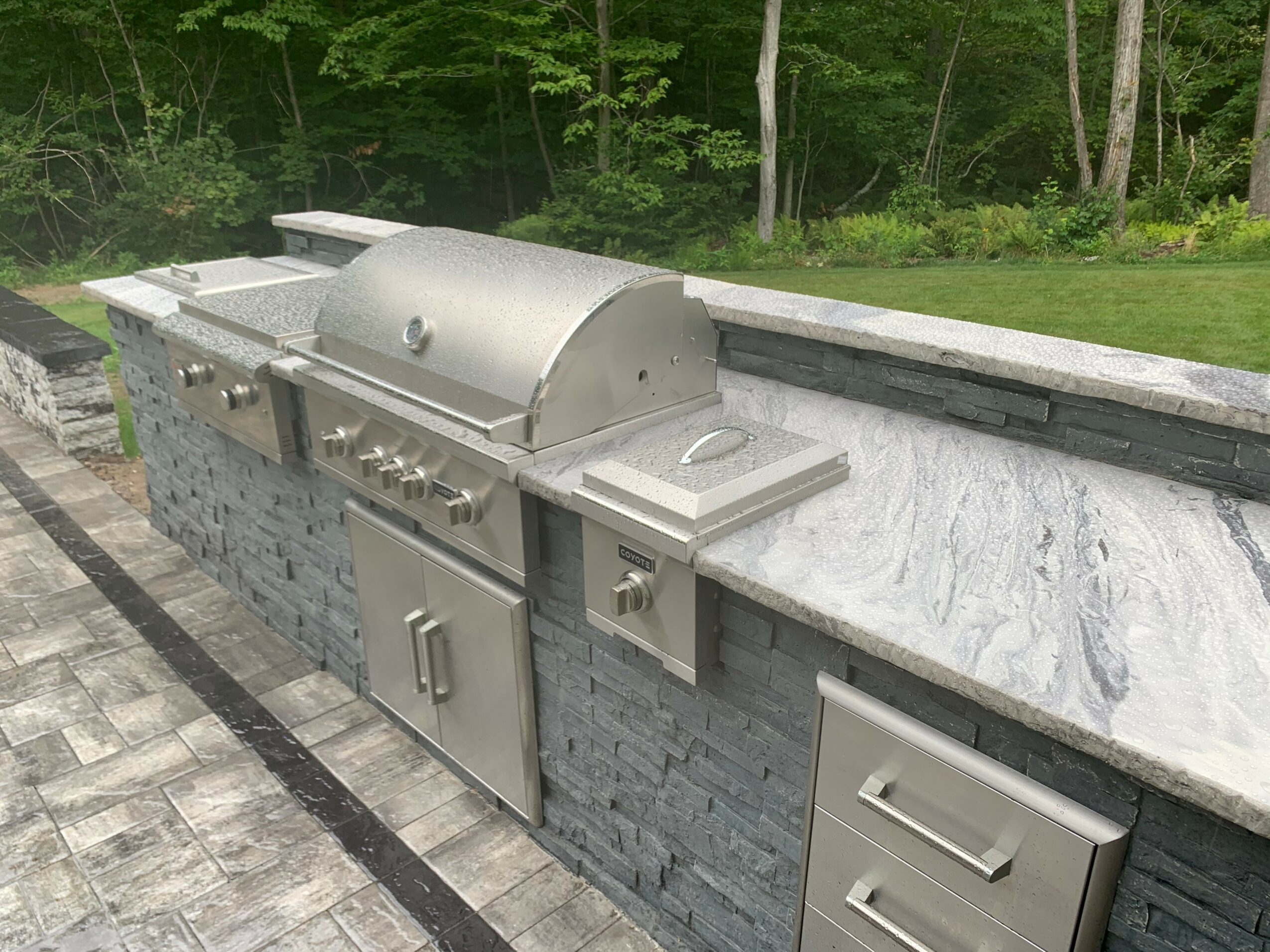 Outdoor stainless steel grill with side burner installed on a stone and marble counter, surrounded by trees and greenery.