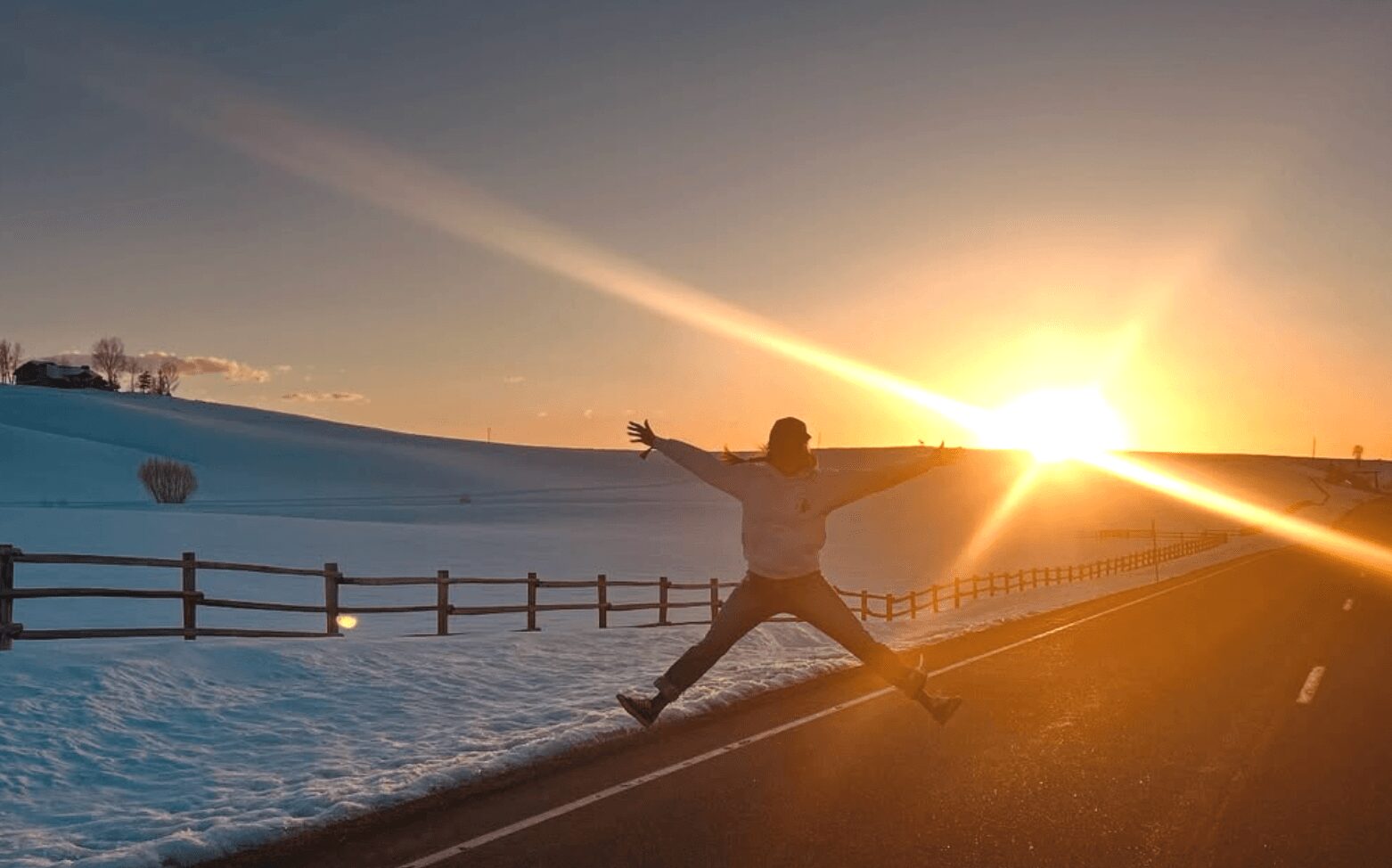Person jumping with arms and legs spread wide on roadside during sunset with snow and fence nearby.