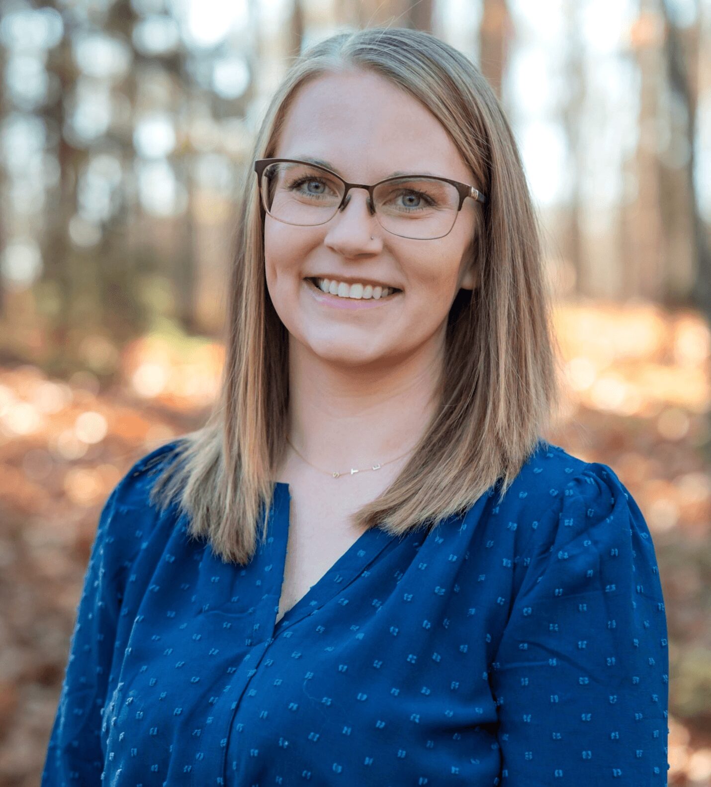 Young woman with glasses smiling outdoors in a forest setting, wearing a blue blouse with small patterns.