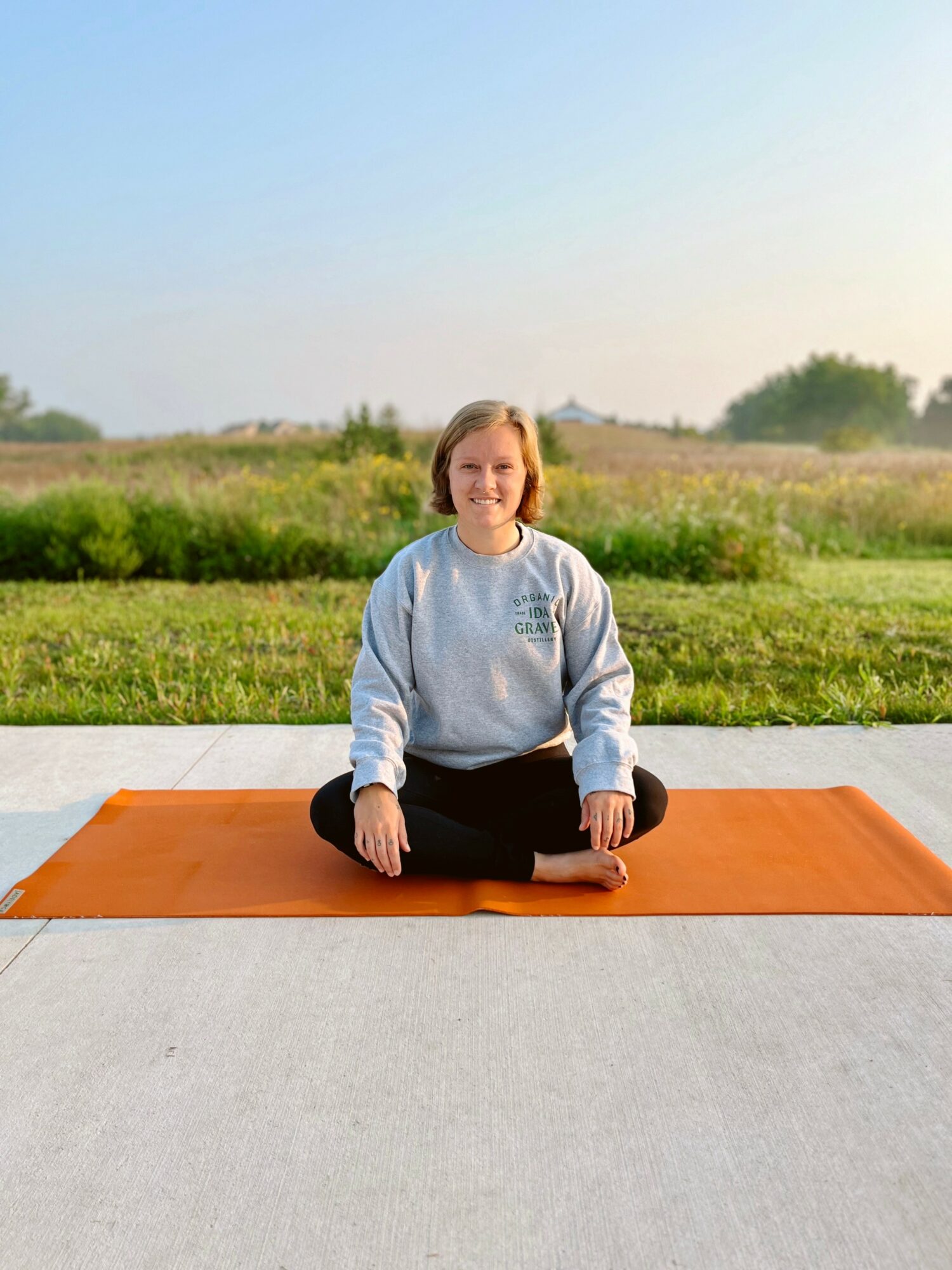 Child sitting cross-legged on an orange yoga mat outdoors, smiling, with green grass and trees in the background.