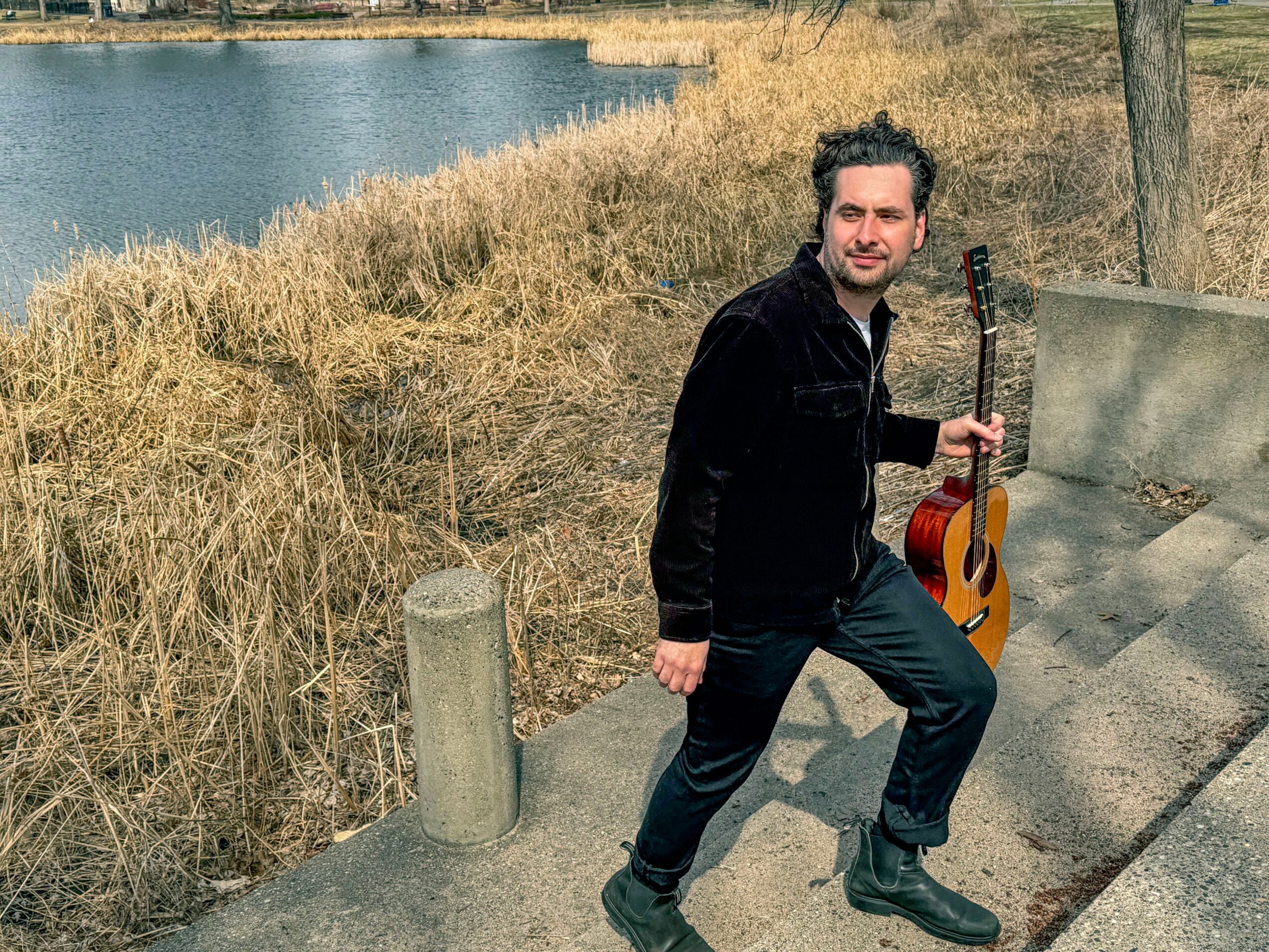 Man standing outdoors near water, holding a guitar, on a paved path with grass and trees.