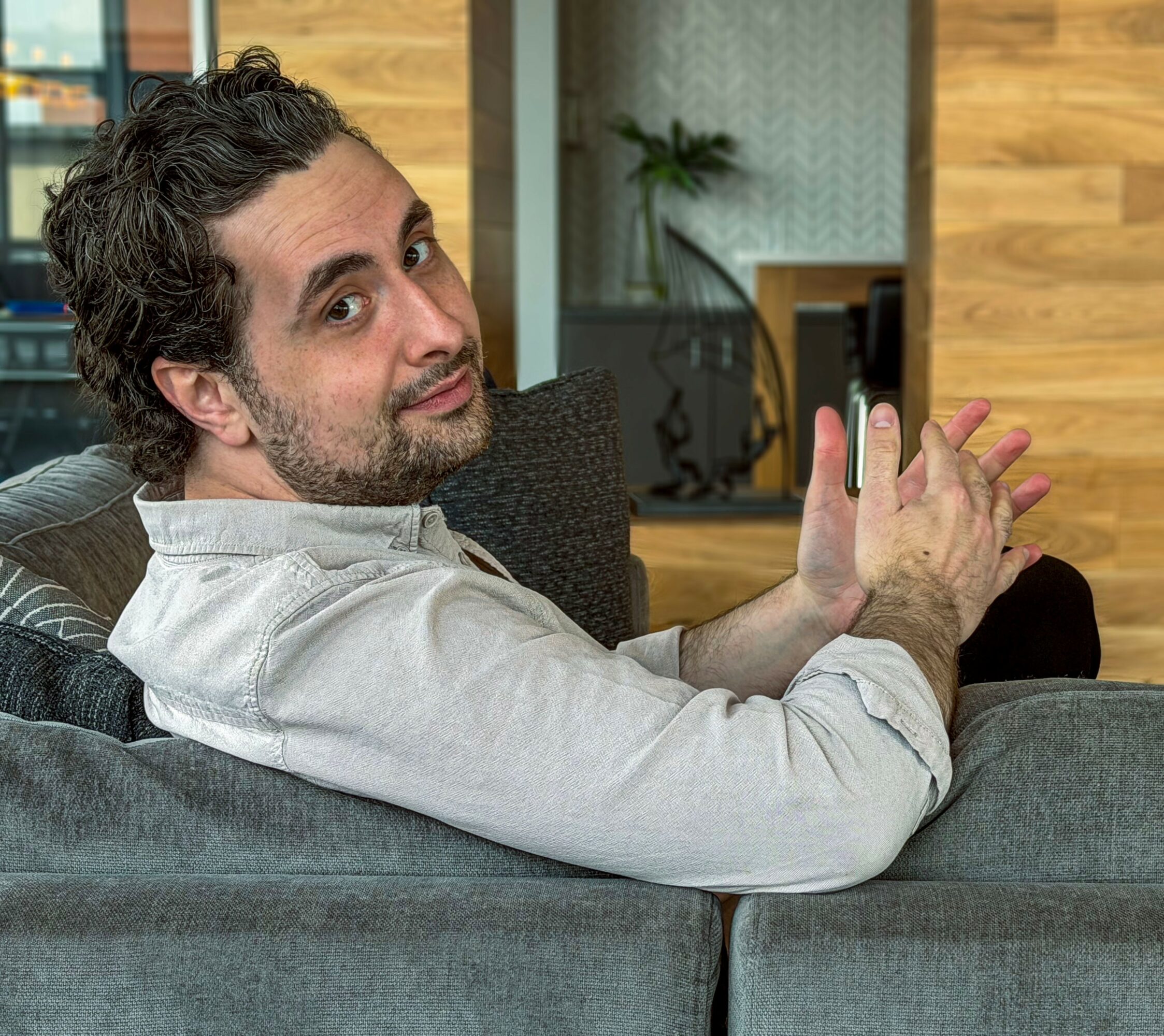 Man with dark curly hair and beard sitting on a sofa, looking at camera with hands clasped.