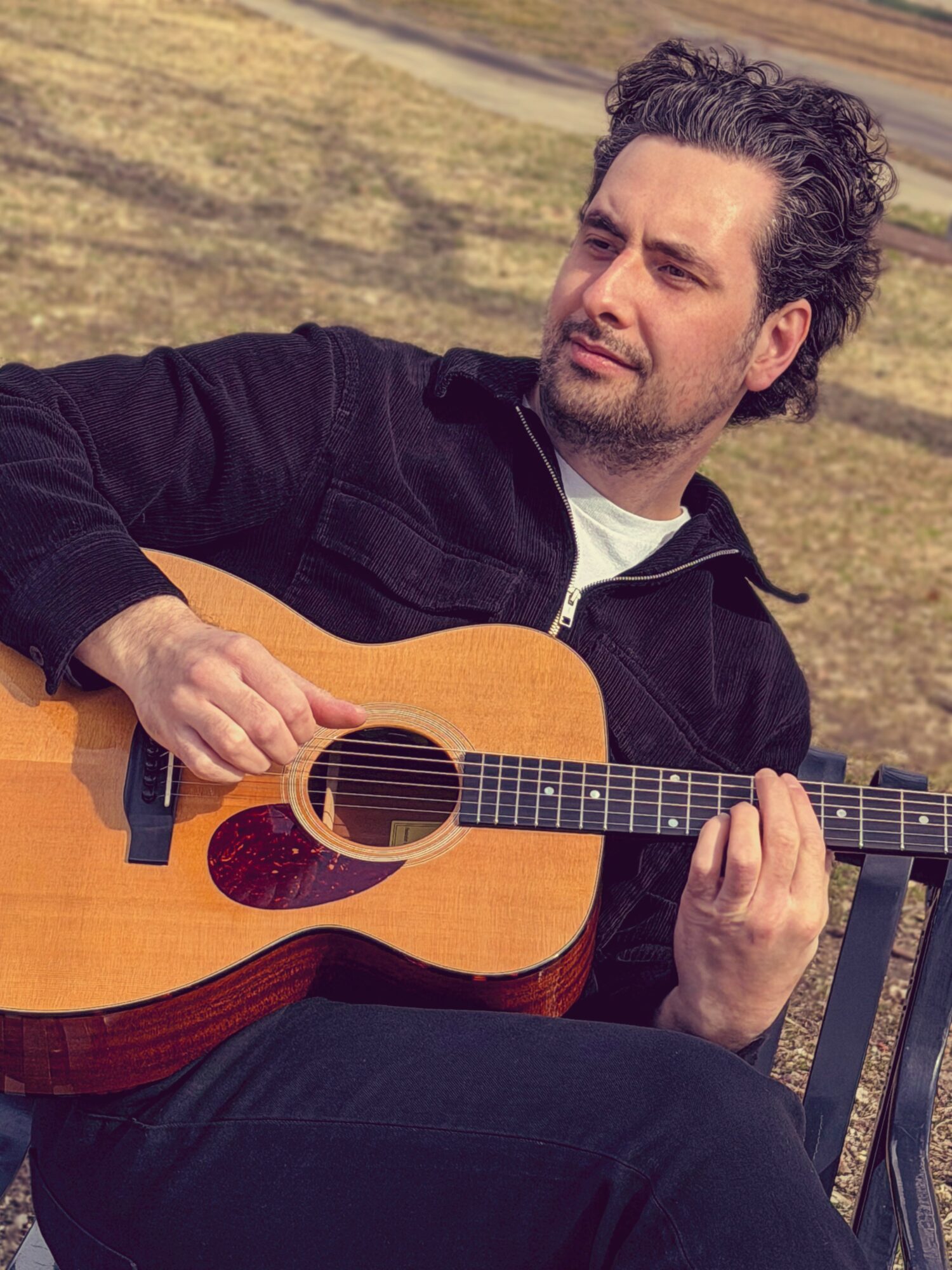 Man with curly hair playing an acoustic guitar outdoors on a bench.