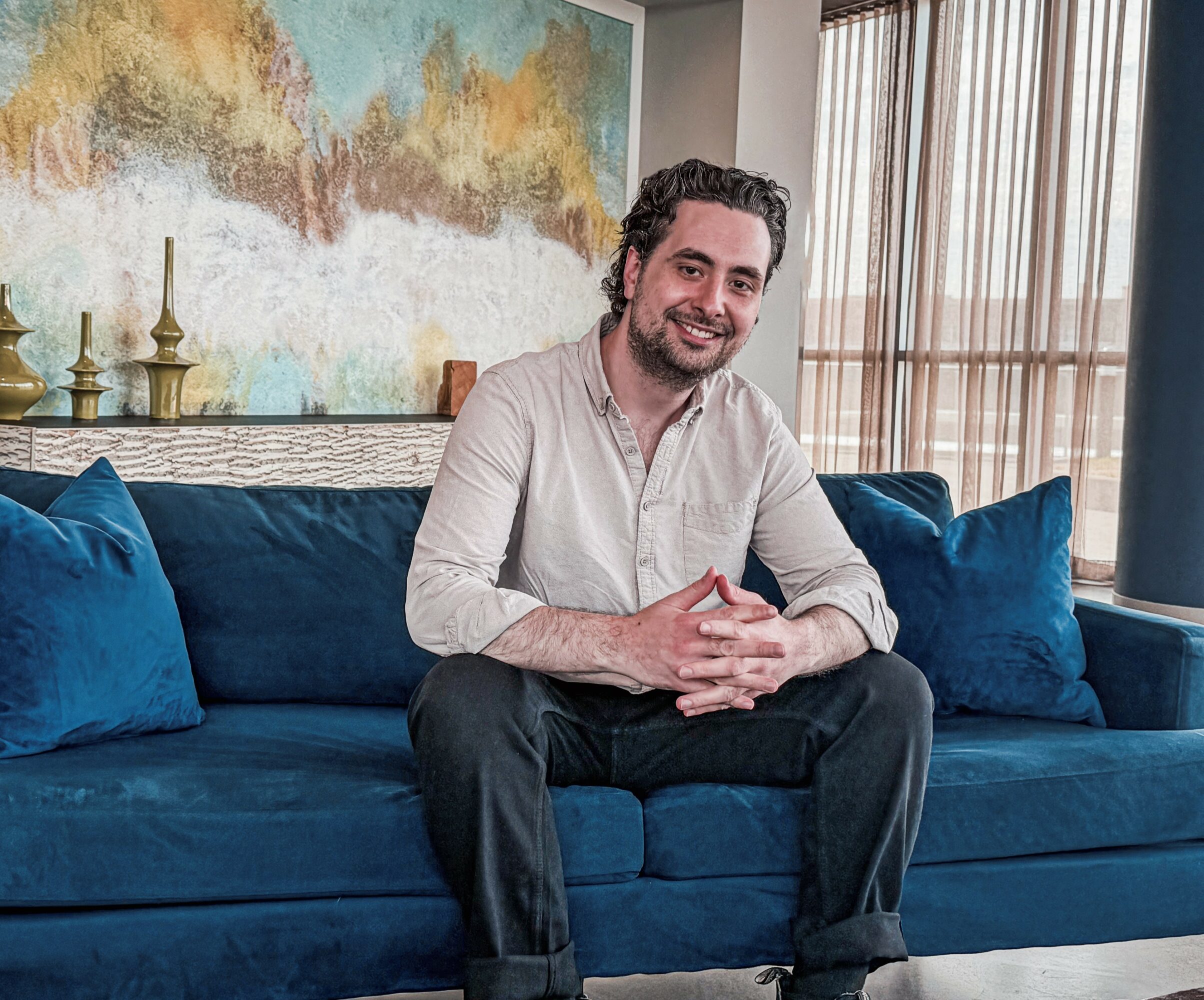 Man sitting on a blue sofa with pillows, smiling, in a room with large windows and artwork on the wall.