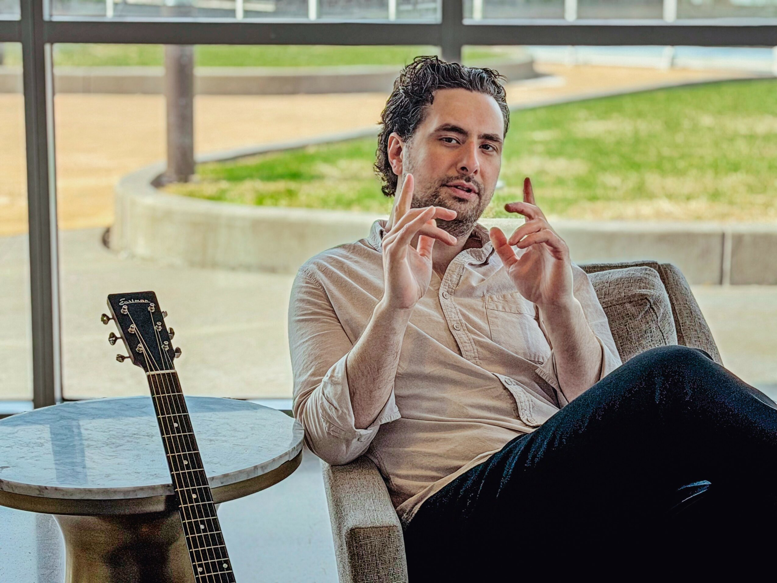 Man sitting on a sofa near a window, gesturing with hands, guitar on a table beside him.