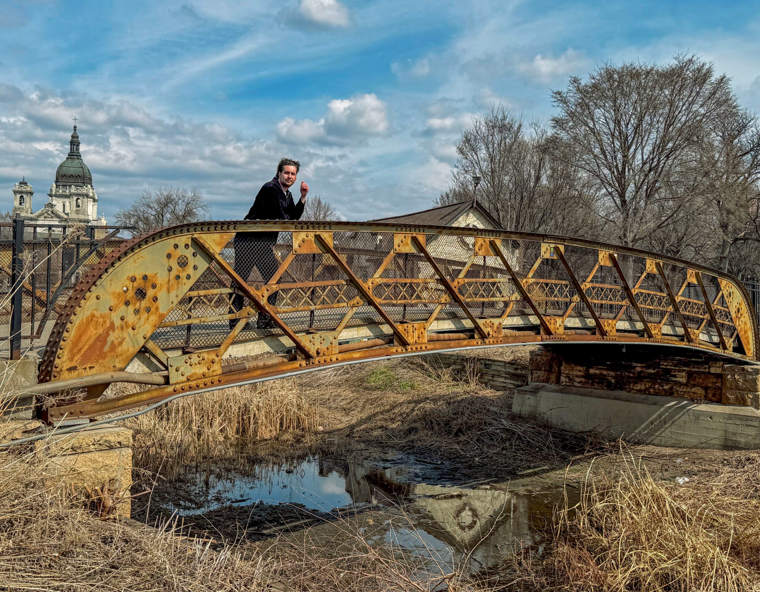 Person standing on a rusty metal bridge over a small waterway with trees and buildings in the background.
