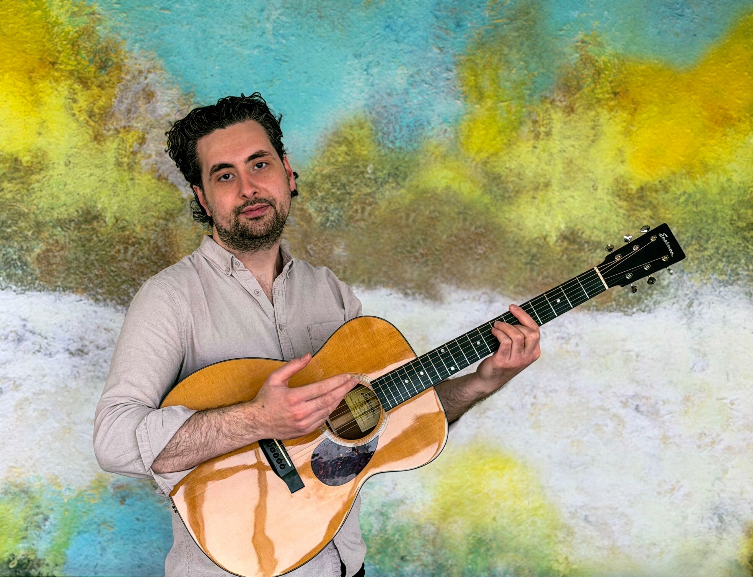 Man holding an acoustic guitar in front of a colorful abstract background.