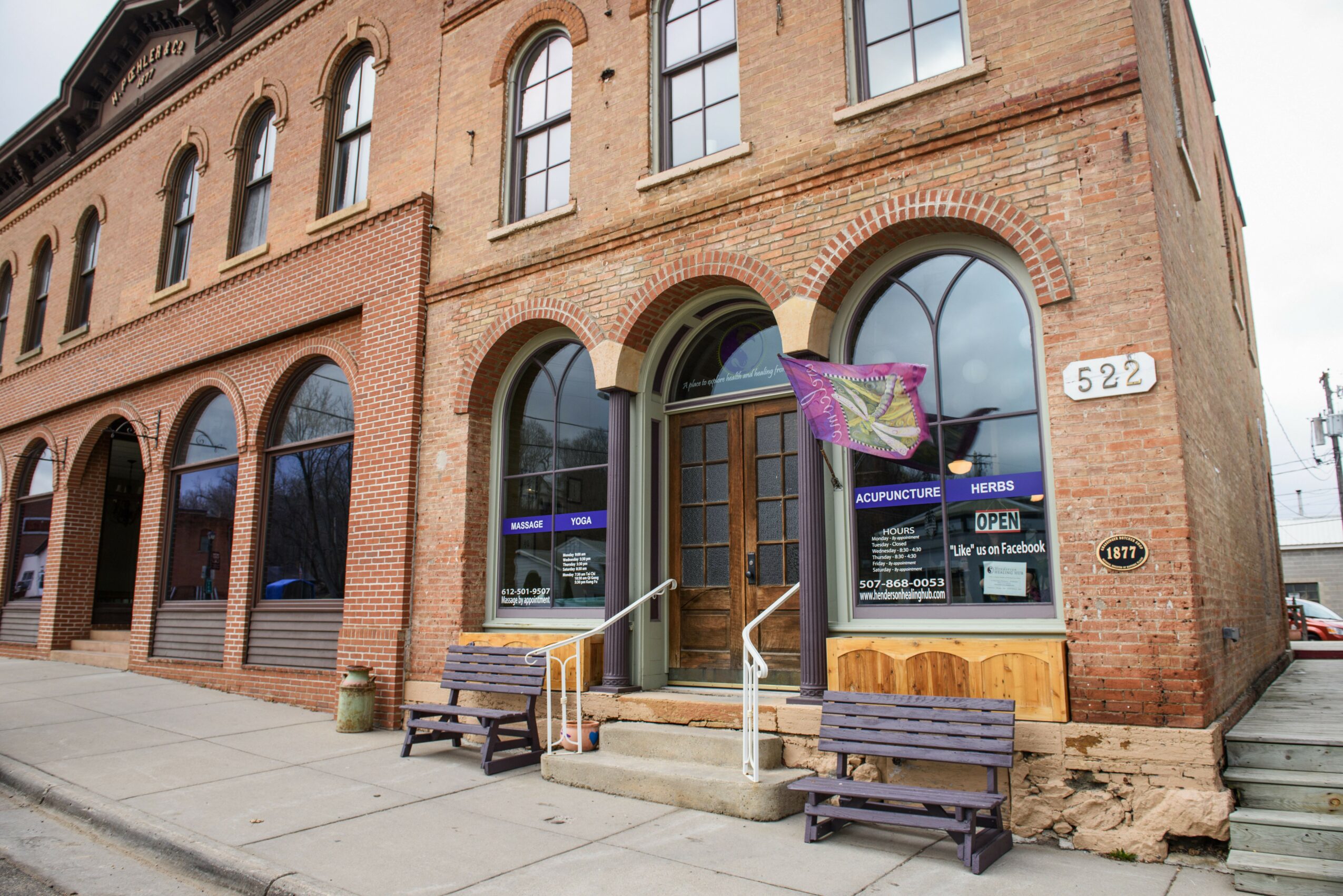 Brick building with large arched windows and a wooden door, two benches outside, and a purple flag near the entrance.