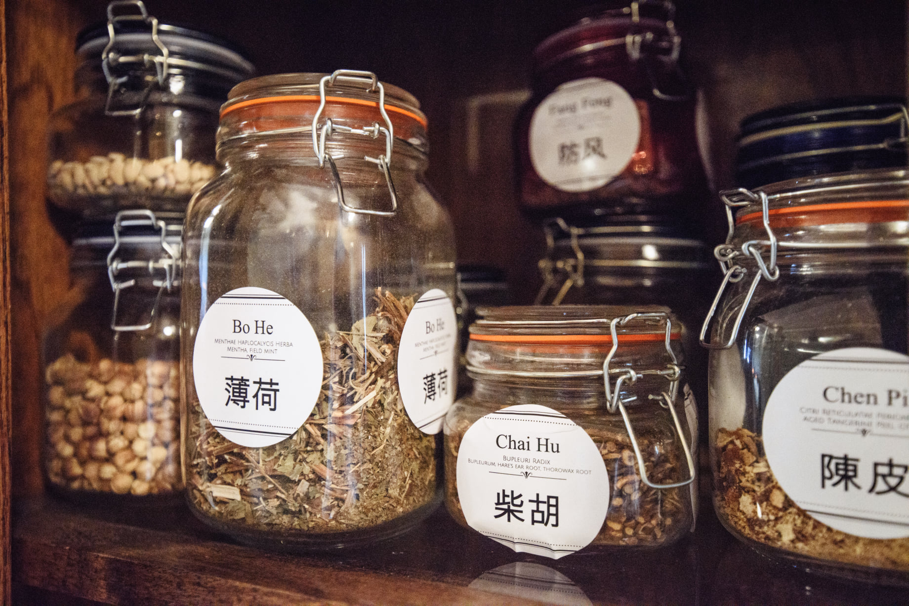 Several glass jars with metal clasps contain dried herbs, seeds, and spices on a wooden shelf.