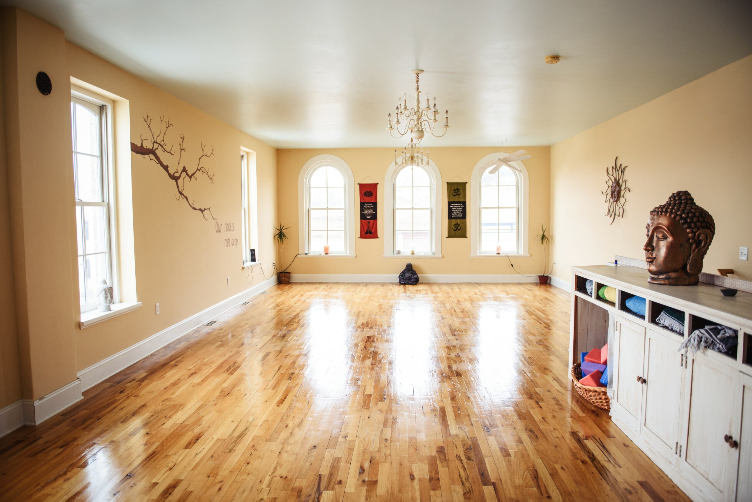 Large, empty room with hardwood floors, three arched windows, a chandelier, and a white cabinet with decorative items.
