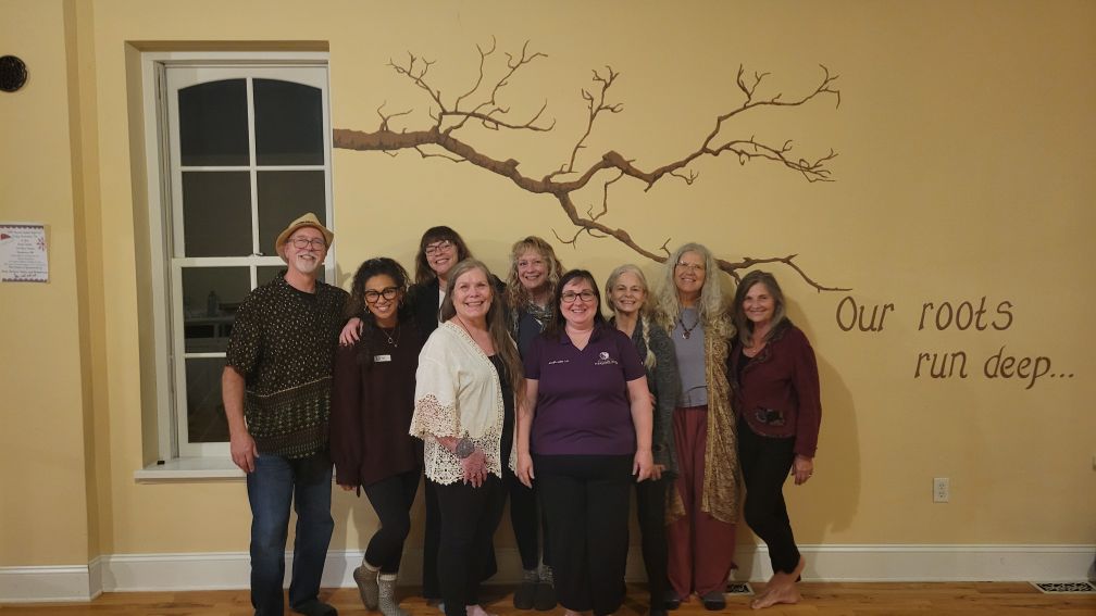 Group of nine women standing indoors in front of a wall with a tree branch mural and text.