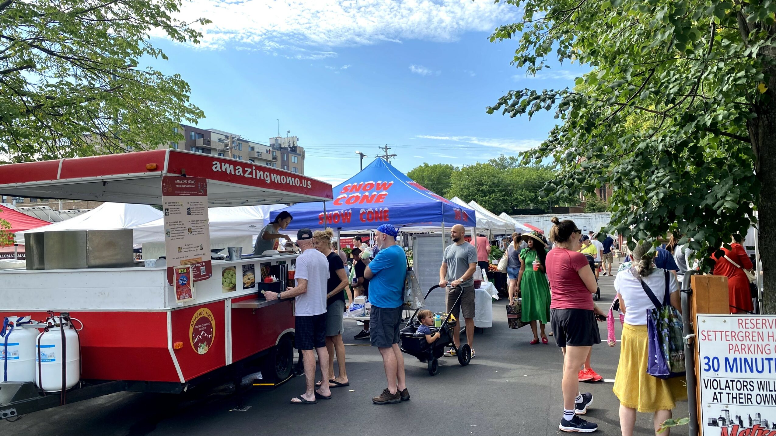 People gather around a red and white food truck at an outdoor event with tents and trees.