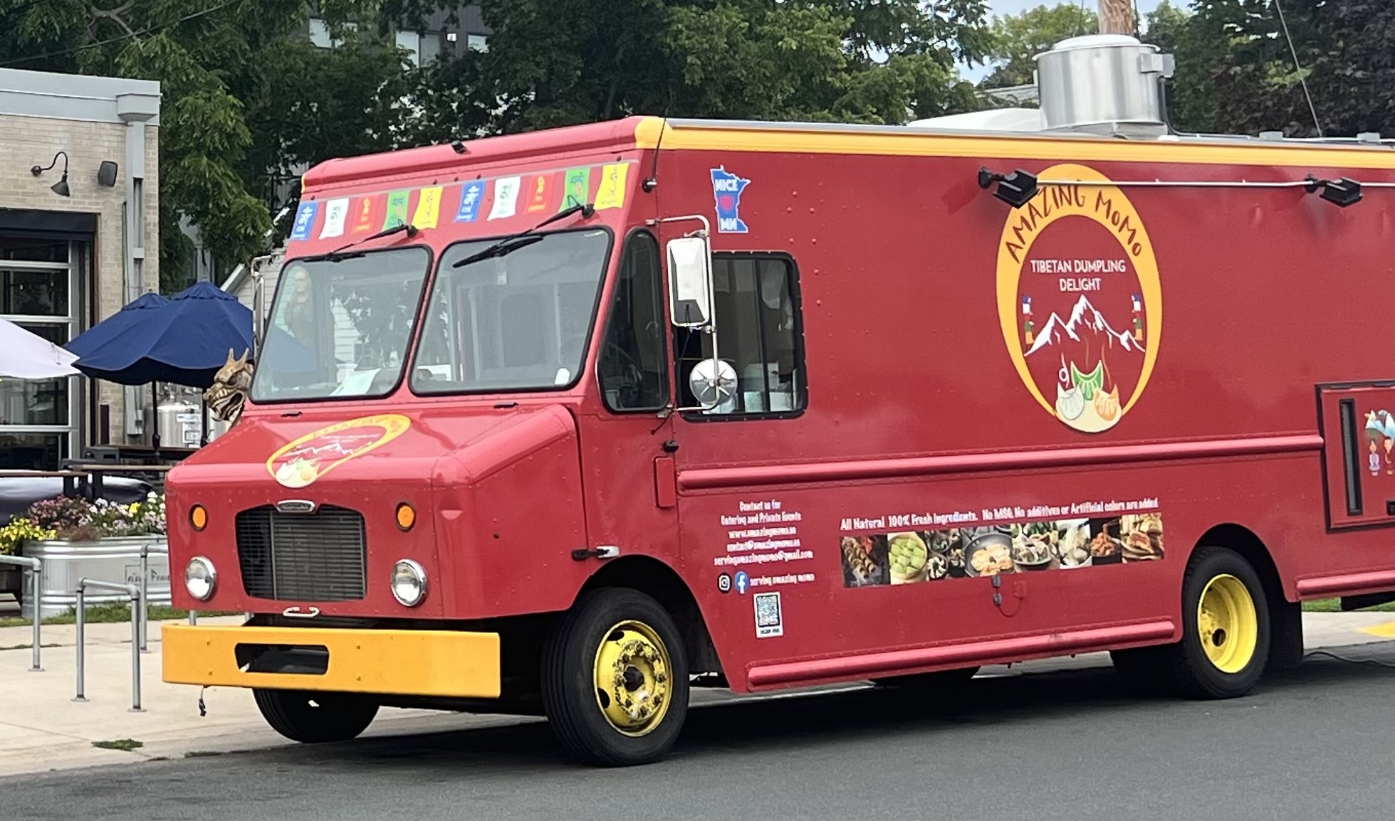 Red food truck with colorful stripes and a logo, parked on street with trees and buildings in background.